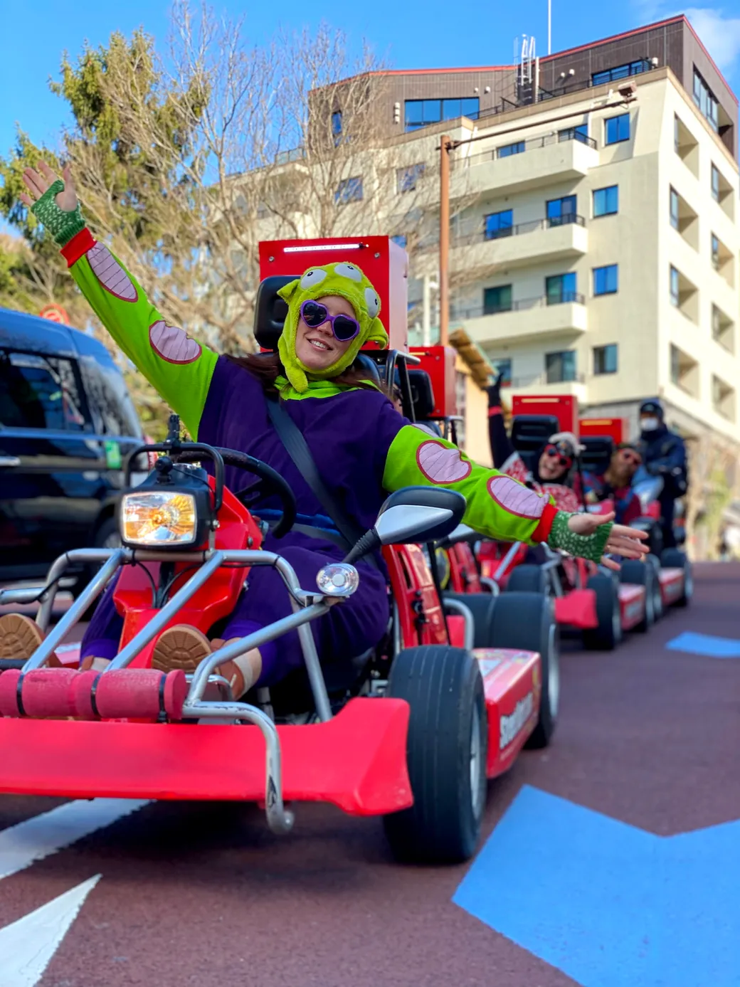 Person in a bright green frog hat and purple costume driving a red go-kart in a street parade, arms raised in joy.