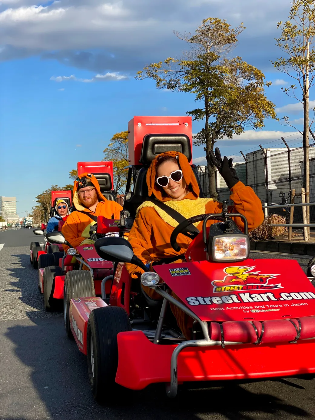 People in orange kangaroo costumes ride red go-karts in a parade, one person waving and wearing heart-shaped sunglasses.